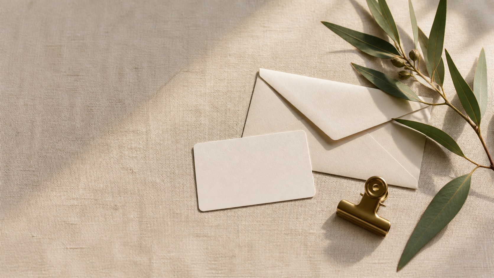A face-down business card, sealed envelope, brass clip and eucalyptus on pale linen.