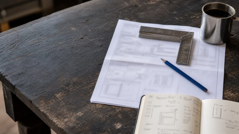 A clipboard and folded plans on a weathered timber table beside a pencil.